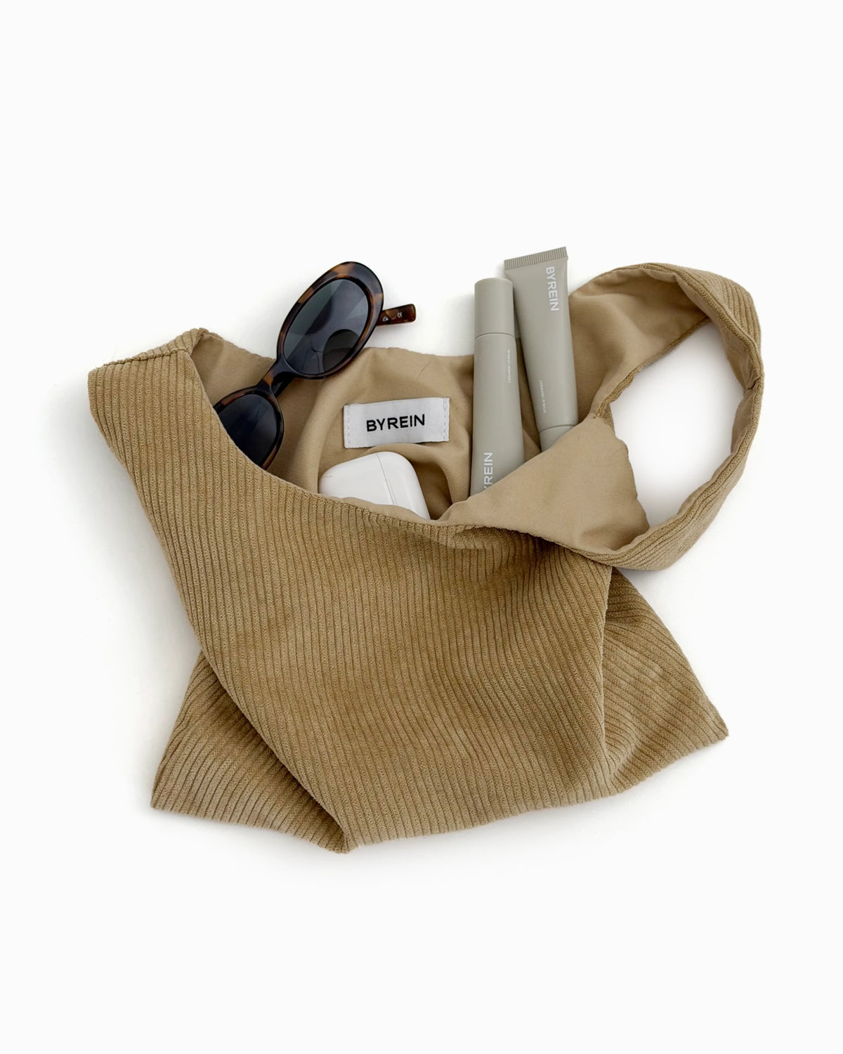 Beige corduroy Hazel Bag laid flat and filled with sunglasses, AirPods and a BYREIN lip balm
